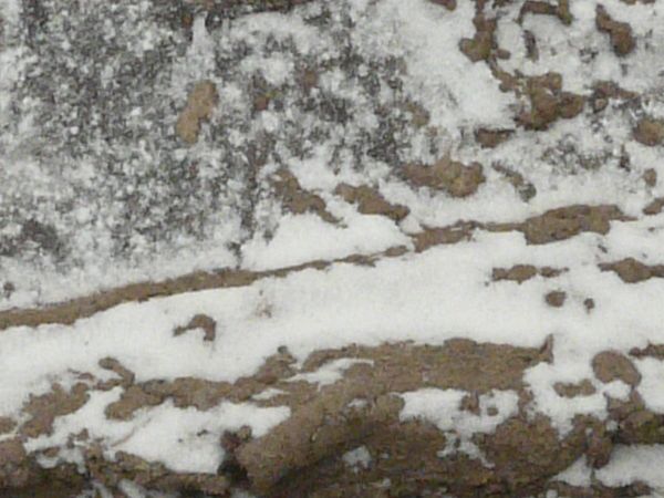 Dirt road texture in dark brown tone with tire marks and thin layer of snow across surface.